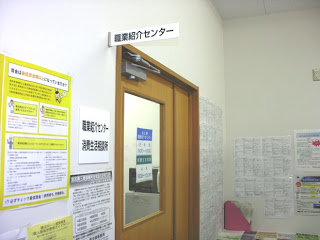 「職業紹介センター」と書かれた看板がドア上と壁に掲示され、周りの壁にはたくさんの掲示物が貼られている「職業紹介センター」の入り口を写した写真