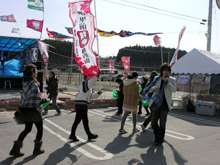 背景に仮設の舞台、Jリーグのフラッグが見える祭り会場で、若者たちが楽しそうに歩いている様子の写真