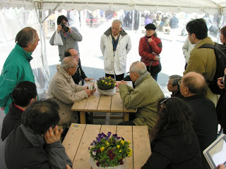 テント内に設置されたテーブルの上に花が飾られている木製のテーブルを囲んで高齢の男性たちが座って話をしている周りにたくさんの人々が集まっている様子の写真