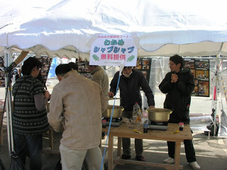「わかめシャブシャブ無料提供」と書かれた看板の下、白いテントの中で数人の男性が鍋で調理し、数名のお客さんが商品を求めて並んでいる写真
