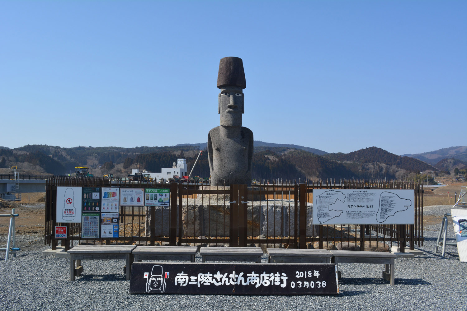 青空の下、柵に囲まれた台座の上に目と帽子がついた大きなモアイ像が立ち、手前にはベンチと黒い看板、そして注意書きや解説板が並んでいる写真