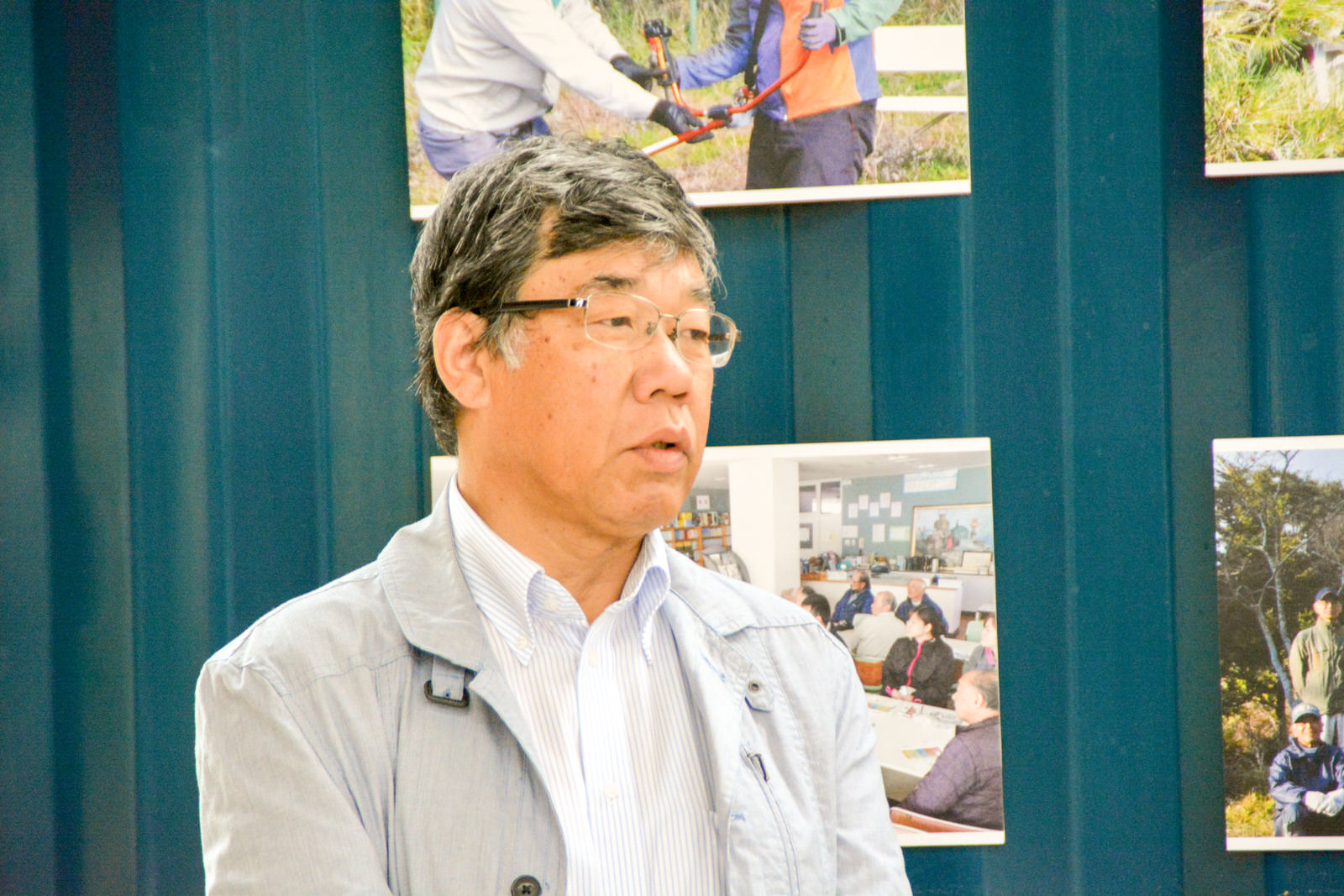 活動の様子を写した写真が展示されている青い壁の前でグレーのジャケットを着た鈴木さんが話をしている写真