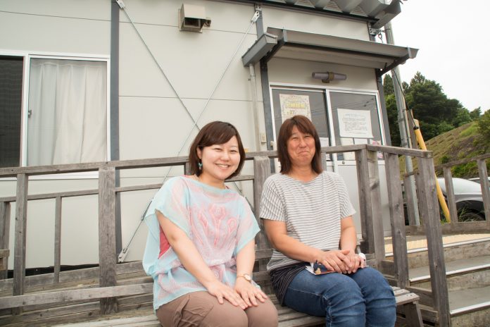 白い壁の建物の、木の手すりの前に置かれたベンチに並んで座っているリポーターの女性と三浦さんの写真