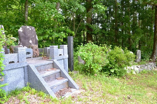 周囲は緑の植物に囲まれ、いくつかの石碑とお墓に上がる小さな階段が自然の中に立っている写真