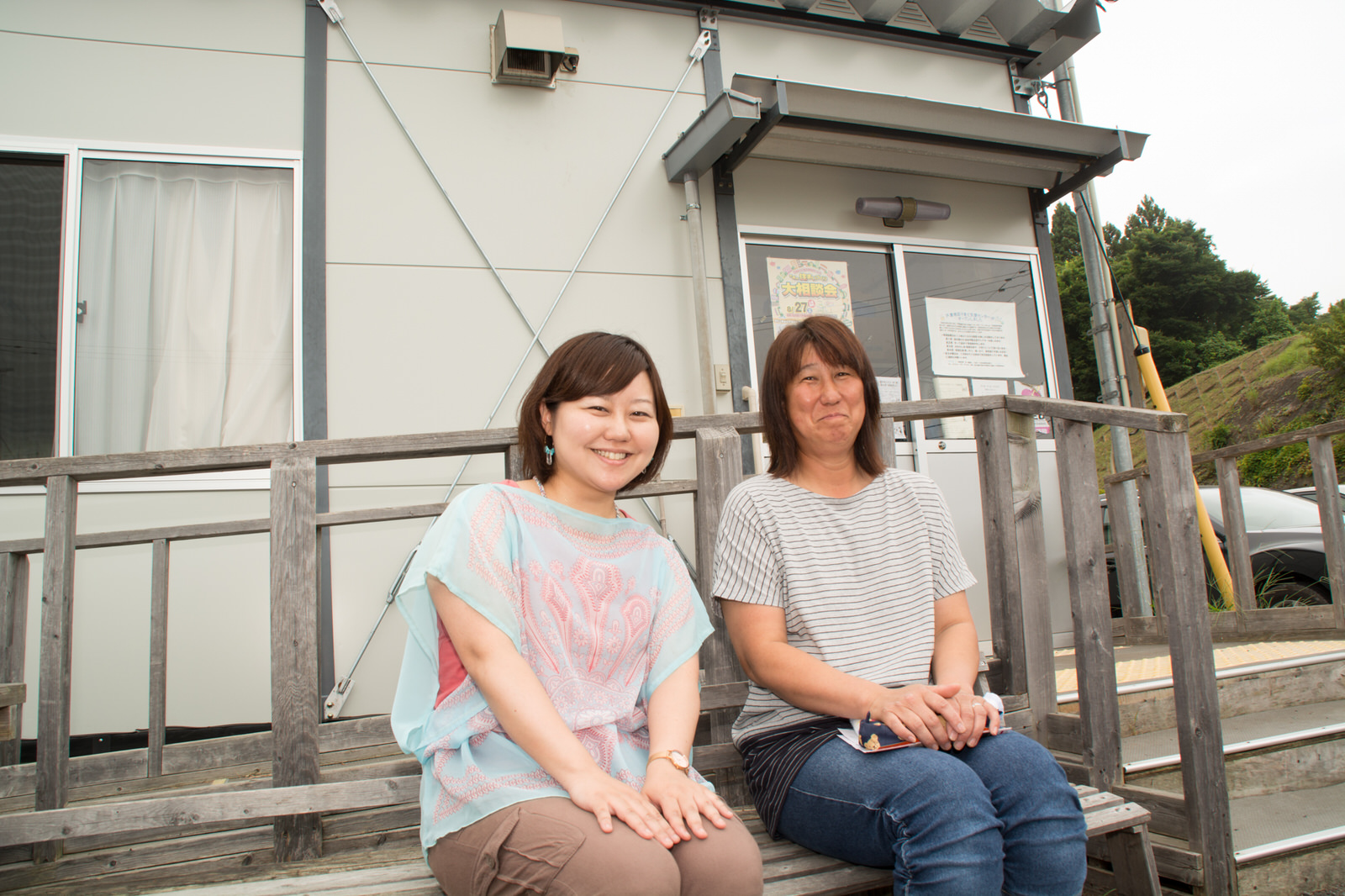 白い壁の建物の、木の手すりの前に置かれたベンチに並んで座っているリポーターの女性と三浦さんの写真