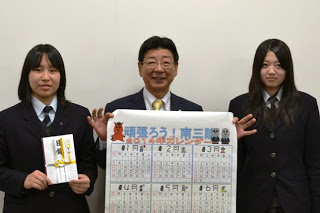 スーツを着た佐藤町長が中央に立ち、両脇には制服姿の女子学生二人が並び、寄付金と来年のモアイカレンダーを持って笑顔で写っている写真