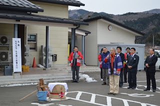 杵と臼が置かれた駐車場で、後ろに赤い法被やスーツを着た人々が立っている中、青い法被を着た男性がマイクを持って話をしている写真