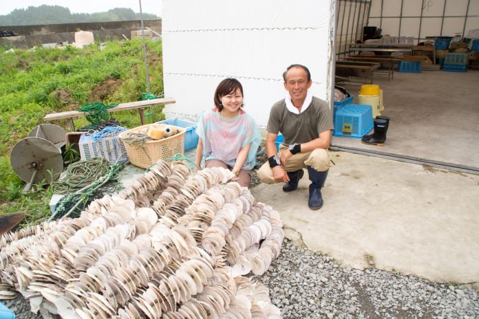 紐を通して束ねた無数の貝殻の山を前に並んでしゃがんでいる村岡さんと女性リポーターの写真