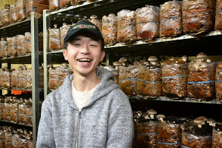 しいたけの菌床が並ぶ栽培棚の前で、灰色のパーカーを着た男性が笑顔で立っている写真