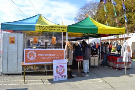 オレンジ色の看板が手前に設置され、衣類などのリユース品が並んだ青・黄色などのテントの下で、買い物を楽しむ人々が集まっている様子の写真