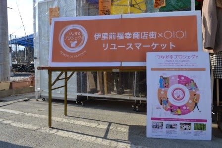 「伊里前福幸商店街×マルイリユースマーケット」と書かれたオレンジ色の大きな案内パネルと、「つながるプロジェクト」を説明するポスターが並んで立てられている写真