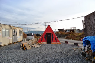 赤と黒で彩られた三角形のピラミッドの建物が中央に立ち、その左右にコンテナハウスの建物やブルーシートで覆われた資材などがある場所を写した写真