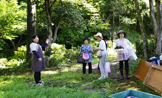 林の中で女性たちがエプロンや帽子を身に着けて集まり、片手にバケツや袋を持ちながら作業の打ち合わせをしている写真