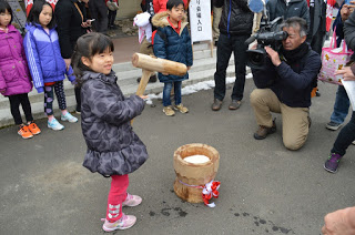 子どもたちが見つめる中、子ども用の杵を持った女の子が、小さい臼の前に立ち、それをビデオカメラを構えた男性が撮影している様子の写真
