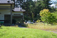 和風の建物の前に芝生と小道があり、その先に軽トラックと数人の人が見える静かな農村風景の写真