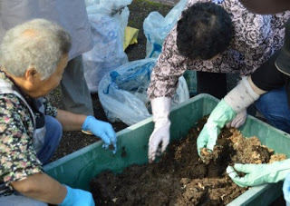 数人の高齢女性が青や白の手袋をして、プラスチック容器に入った土を混ぜながら作業している写真