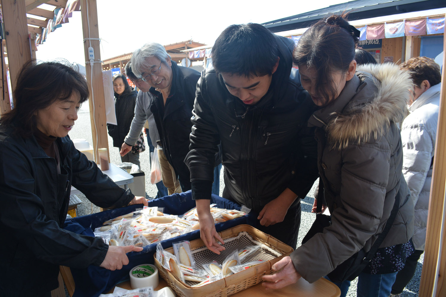 屋台の店舗前で、来場者が箱にたくさん入った地元の名物である笹かまぼこを手に取りながら店員の説明を受けている様子を写した写真