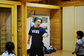 木の温もりを感じる室内空間で、深緑色のヘッドバンドを着けた若い男性が子どもたちに向かって何かを楽しげに話しかけている様子をを写した写真