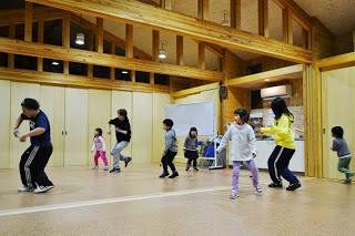 木造の温かみある室内で、カジュアルな服装の複数の子どもたちが左から右へ1列に並び、インストラクターに合わせて同じ動きをしている様子を写した写真