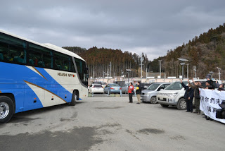 曇り空の下、駐車場から出発する白と青の配色の大型バスに向かって、集まった人々が手を振り、見送っている様子の写真