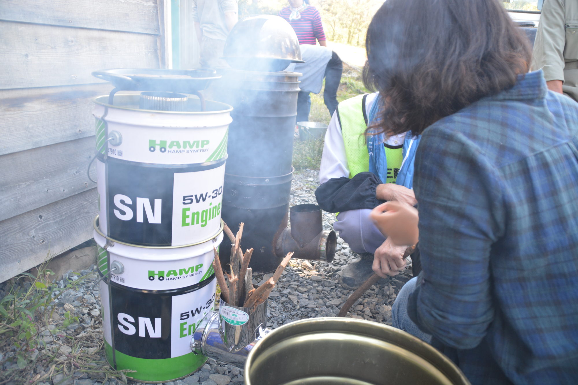 屋外で複数の人が木製の構造物のそばに集まり、オイル缶で作られたロケットストーブから煙が立ち上る中、チェック柄のシャツを着た女性が棒や道具を使って火や加熱作業をしている様子が写された写真