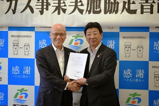 南三陸町のバックパネルの前で、スーツ姿の男性と佐藤町長が協定書を手に笑顔で握手を交わしている記念写真