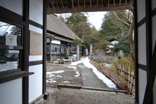 白壁の建物の門の先に雪の残る道が延び、左側に茅葺屋根の建物が建っている写真