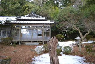 雪が積もった植栽と石灯籠がある庭の奥に、瓦屋根とガラス戸の伝統的な日本家屋が建っている写真