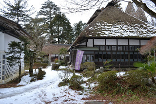 雪が積もった道の左側に白壁の建物、右側に藤色ののぼりがあり、その後ろに茅葺き屋根の伝統的な日本家屋が建っている写真
