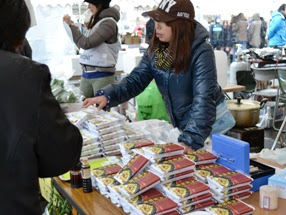 屋外の出店で、「めかぶ納豆」パッケージ商品が多数並べられており、女性の店員が販売している様子を写した写真