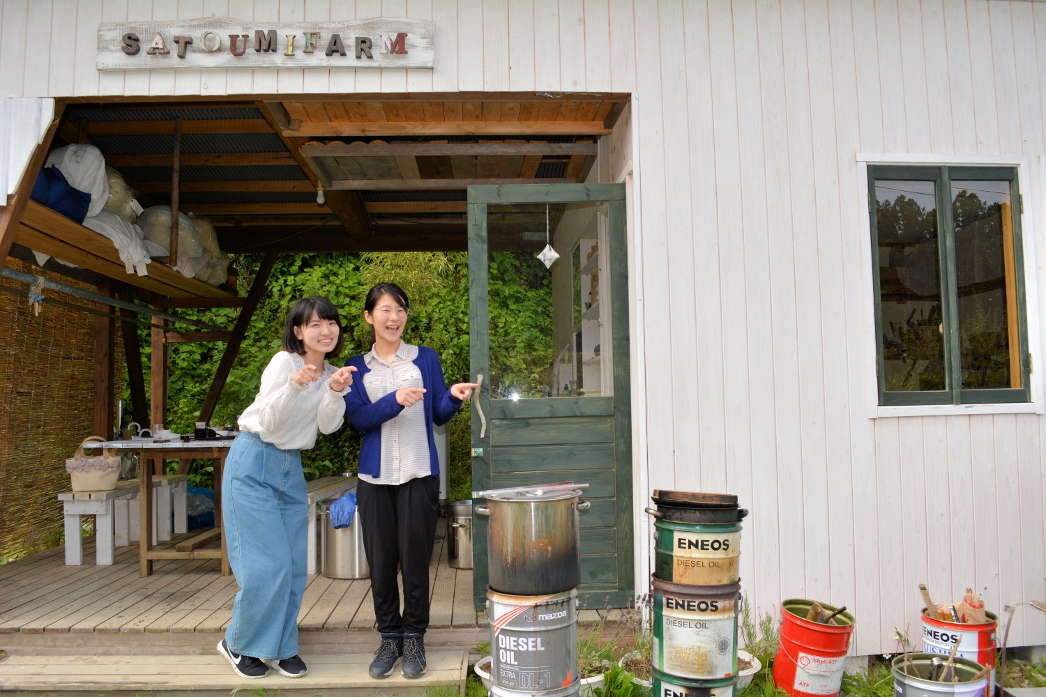 「SATOUMIFARM」と書かれた木製の看板が取り付けられた白い壁の建物の前で、2人の女性が笑顔で左を指を差してポーズをとっている写真