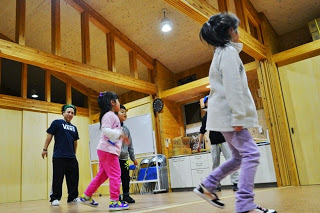 一番手前にいる女の子は、白い長袖とラベンダー色のズボンを着用し、横を向きながらステップを踏んでいるような姿で、中央の女の子はピンクのズボンと白いTシャツ姿で、リズムに乗って動き奥には指導者と思われる男性が、見守るように立っている室内写真