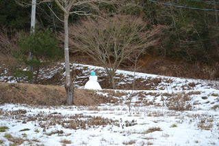 雪が残る斜面の木々の間に、帽子に見立てた水色のバケツを頭に乗せた雪だるまの後ろ姿が見える写真
