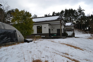 木々に囲まれた、瓦屋根に雪が積もった平屋のシルク館の外観写真