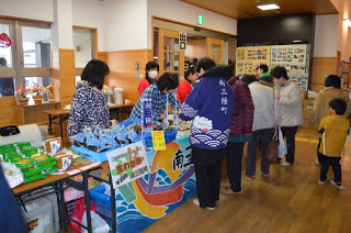 南三陸町の販売ブースのテーブルの上に生わかめなどの商品がたくさん並べられ、紺色の法被を着た男性や、販売係の女性たちが、訪れた女性客らに接客している様子の写真