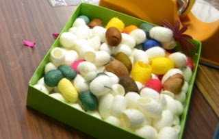 緑色の正方形の箱の中に、白、茶色、黄色、緑、ピンクなどの繭がたくさん入っている写真