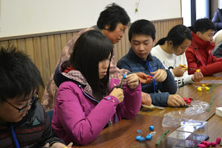 テーブルに並んで座った子どもたちが、思い思いの色の繭を使って制作に取り組み、後ろから女性が指導をしている様子の写真