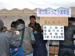 南三陸ふっこう青年会のテントの下で、数人の来場者が列を作り、「南三陸産塩蔵ワカメ詰め放題1袋1000円」と書かれた手作りの看板の前で、笑顔の男性が接客している出店の様子を写した写真