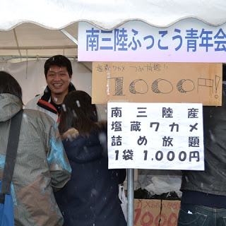 南三陸ふっこう青年会の看板があるテントの下で笑顔の男性が接客しており、ワカメを販売している様子がわかる写真