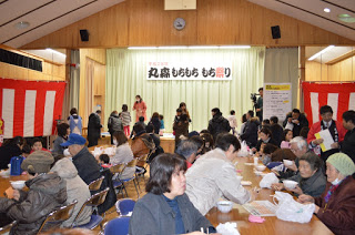 「丸森もちもち もち祭り」の横断幕が掲げられたステージの前にたくさんの人が集まり、会場に並べられたテーブルの席も大勢の来場者で埋まっている会場の様子の写真