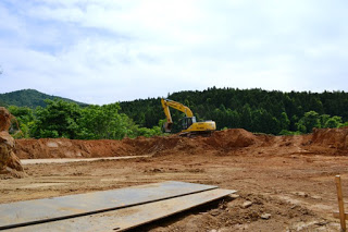 掘削された赤土と鉄板が置かれた広い工事現場で、黄色い油圧ショベルが土の盛り上がった場所で稼働している様子を写した写真