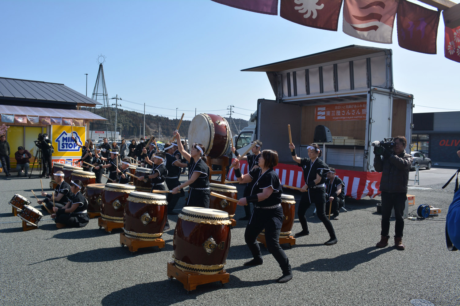 黒い衣装を着た大森創作太鼓の皆さんが商店街前の広場で力強く和太鼓を演奏している様子を写した写真
