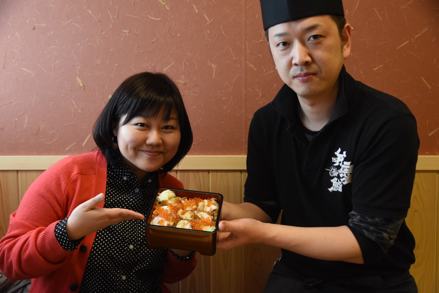 イクラなどの海鮮が盛り付けられた重箱を女性リポーターと黒い制服を着た菅原さんが一緒に持って笑顔を見せている写真