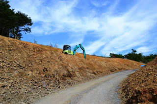 右にカーブしながら続く坂道の左側で緑色のショベルカーが作業をしている様子を写した写真