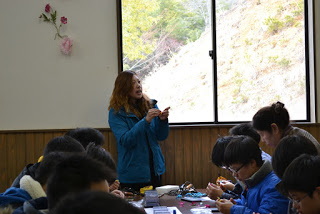 子どもたちがまゆ細工をしているテーブルの横に立った通訳の女性が話をしている写真