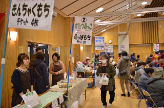 「きんちゃくもち チケット4枚」などののぼりを掲げた長机に商品が並び、パイプ椅子が並んだ会場が大勢の来場者でにぎわっている様子の写真