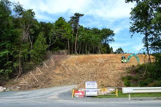 土がむき出しになっている造成工事が始まった斜面に重機が見え、手前には工事案内の看板が見えている工事現場の写真