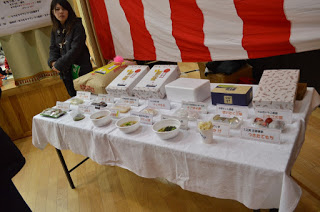 紅白幕の前に置かれた白いクロスが掛かったテーブルの上に、出品している料理の数々と、賞品の米や包装された箱などが並べて置かれ、左側に女性が一人立っている写真
