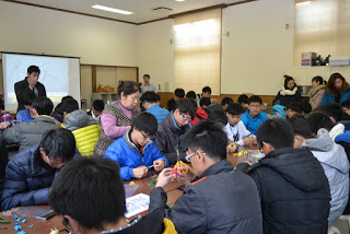 会場の2列のテーブルに向かい合って座った子どもたちが、まゆ細工に夢中で取り組んでいる様子の写真