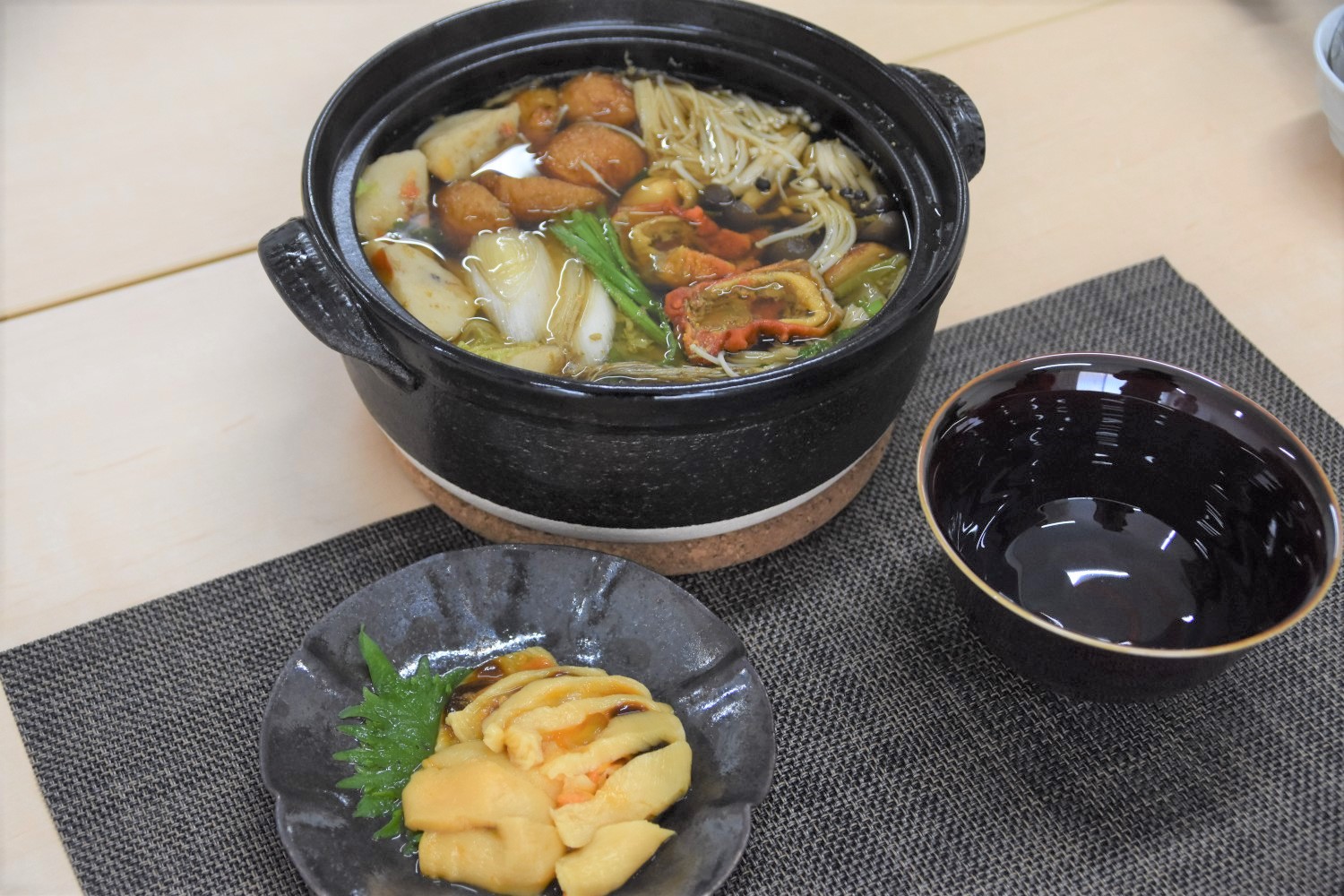 黒い土鍋にきのこや野菜、練り物が入った汁物と、その前にホヤが入った小皿と黒い汁椀が並べられているテーブル上の料理の写真